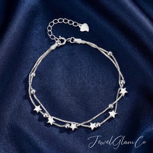 May include: A silver bracelet with two strands, adorned with small star charms and round beads. It has an adjustable chain with a heart-shaped charm. The background is a dark blue fabric. The text "JewelGlam Co" is in the bottom right corner.