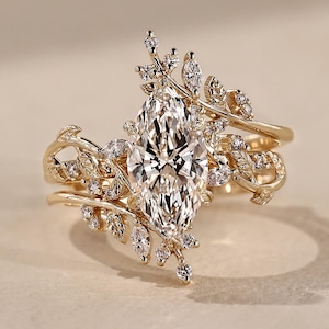 Könnte beinhalten: Ein goldener Ring mit einem großen, marquise-geschliffenen Mittelstein, der von kleineren marquise- und rundgeschliffenen Steinen umgeben ist. Die Ringschiene ist mit filigranen Goldblattmotiven verziert, die nach außen ragen und ein detailliertes, organisches Design bilden.