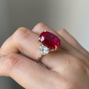 Könnte beinhalten: Ein großer, oval geschliffener Rubinring mit Goldfassung und Silberband. Der leuchtend rote Rubin wird von zwei dreieckig geschliffenen Diamanten flankiert. Der Ring wird auf einer Hand präsentiert, mit dem Text "sunshin jewels".
