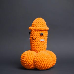 May include: A handmade, orange crochet sculpture of a smiling figure with a hat and two round shapes below. The sculpture has black eyes and a white rectangle for a mouth. The background is a solid grey.