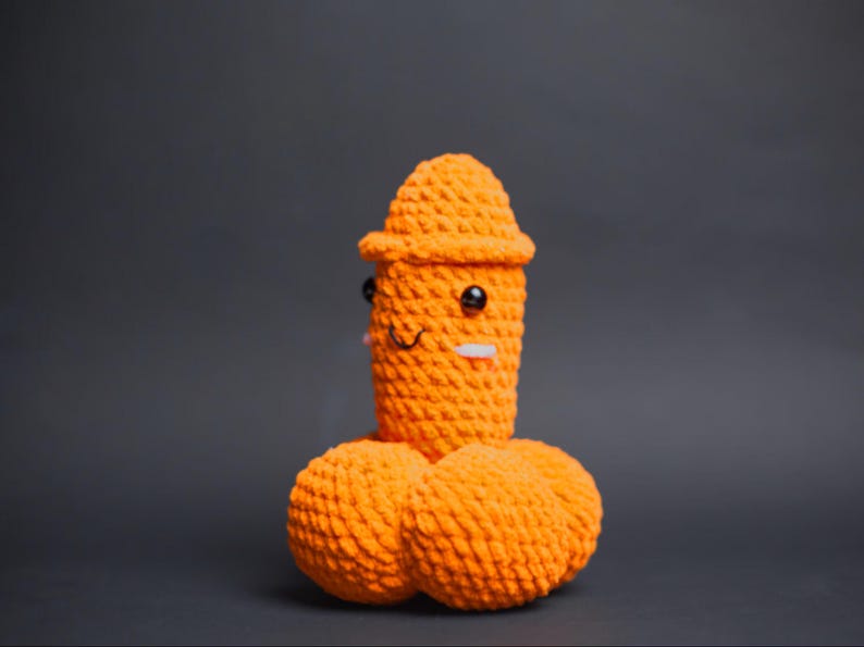 May include: A handmade, orange crochet sculpture of a smiling figure with a hat and two round shapes below. The sculpture has black eyes and a white rectangle for a mouth. The background is a solid grey.