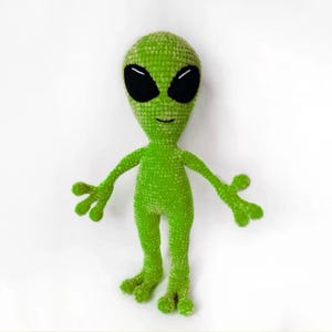 May include: A handmade green crochet alien doll with large black eyes and small arms and legs. The alien has a textured surface and is standing upright. The doll is a whimsical, unique item.