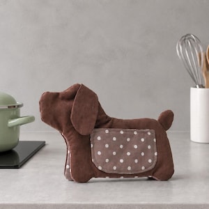 May include: A brown fabric dog-shaped oven mitt featuring a side pocket with a white polka dot pattern. The plush, stuffed animal-like kitchen accessory has a rounded head, floppy ears, and a small tail, designed for handling hot cookware items.
