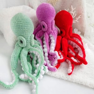 Puede incluir: Tres pulpos de ganchillo en verde, morado y rojo. Cada pulpo tiene múltiples tentáculos con detalles blancos. Los juguetes se exhiben sobre una superficie blanca y texturizada.
