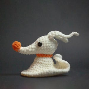 Puede incluir: Una figura de ganchillo hecha a mano de Zero, un perro fantasma de Pesadilla antes de Navidad. La figura es blanca con una nariz y un collar naranjas, y ojos negros. El perro tiene una nariz larga y curvada y orejas caídas.
