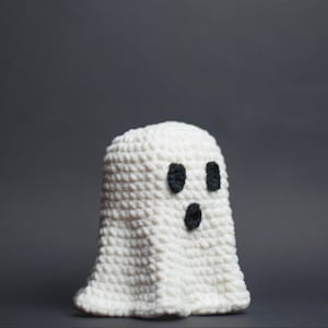 May include: A handmade, white crochet ghost with three black facial features. The ghost has a rounded top and a flowing bottom, ideal for Halloween decorations or themed gifts. The ghost is set against a grey background.