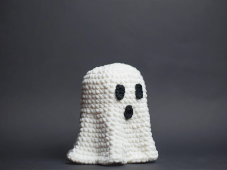 May include: A handmade, white crochet ghost with three black facial features. The ghost has a rounded top and a flowing bottom, ideal for Halloween decorations or themed gifts. The ghost is set against a grey background.