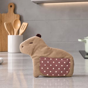 以下が含まれることがあります： カピバラの形をしたベージュ色の布製キッチンアクセサリー。頭部には小さなダークブラウンの房があり、側面には白い水玉模様のマルーン色のポケットが付いています。刺繍された目と鼻があり、柔らかい質感の見た目をしています。