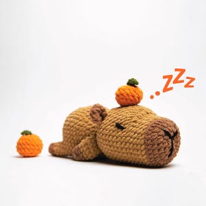 Peut inclure: Une peluche capybara au crochet, en position de sommeil, avec une petite citrouille orange sur la tête et une autre à côté. Le mot "Zzz" est écrit en orange. Le capybara est marron et beige.