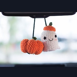 May include: Crocheted Halloween car charm featuring a pair of orange pumpkins and a white ghost with an orange hat. The handmade decorations are suspended from a black string, perfect for seasonal decor.