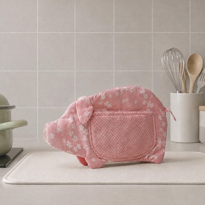 Könnte beinhalten: Ein rosafarbener Ofenhandschuh aus Stoff in Schweineform mit weißem Blumenmuster auf dem Körper und weißem Punktemuster auf der Seitentasche. Das Schwein hat ein kleines Ohr, einen Rüssel und einen kurzen Schwanz aus gestepptem Textil.