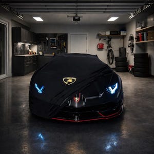 Puede incluir: Un coche deportivo negro parcialmente cubierto, revelando el frente con faros azules. El coche tiene una línea roja y un emblema dorado. La imagen está tomada dentro de un garaje.