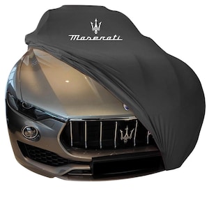 Puede incluir: Una funda negra para coche con el logotipo de Maserati en blanco. La funda cubre un Maserati gris, ocultando parcialmente la parte delantera del vehículo. El emblema de Maserati es visible en la parrilla.