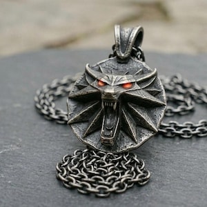 May include: A silver pendant necklace with a wolf's head emblem and glowing red eyes. The pendant is attached to a chain necklace. The wolf head is set within a circular frame with a starburst design.