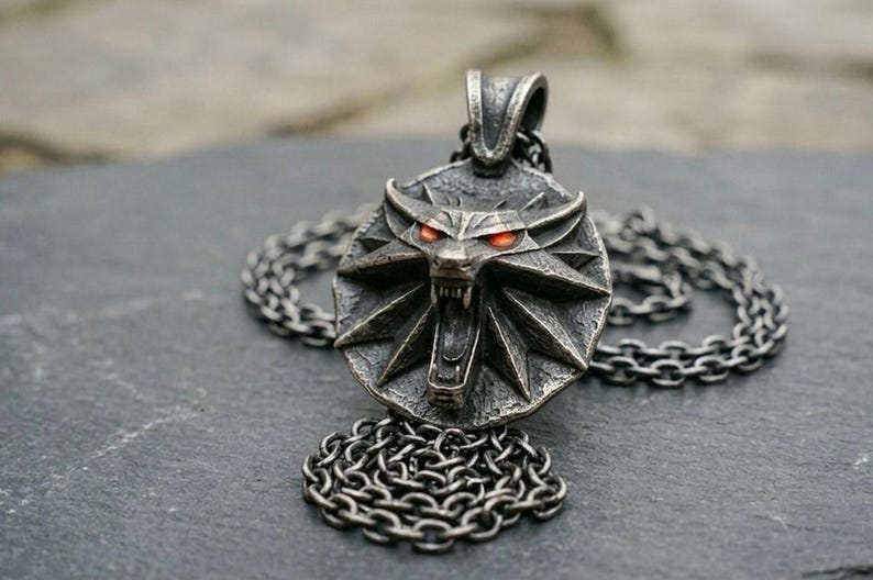 May include: A silver pendant necklace with a wolf's head emblem and glowing red eyes. The pendant is attached to a chain necklace. The wolf head is set within a circular frame with a starburst design.