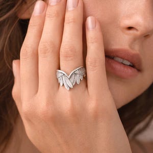 May include: A silver-colored ring with a detailed wing design. The ring features a central feather-like element flanked by two wings, creating a symmetrical and elegant appearance. The ring is worn on a finger.