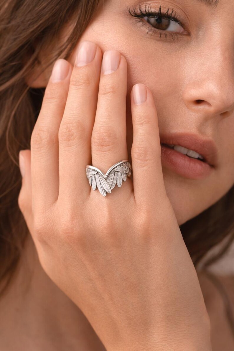 May include: A silver-colored ring with a detailed wing design. The ring features a central feather-like element flanked by two wings, creating a symmetrical and elegant appearance. The ring is worn on a finger.
