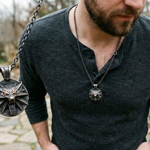 May include: A silver pendant necklace with a wolf's head emblem and red eyes. The circular pendant has a detailed design. The necklace is worn over a dark grey long-sleeved shirt. The background is a blurred outdoor scene.