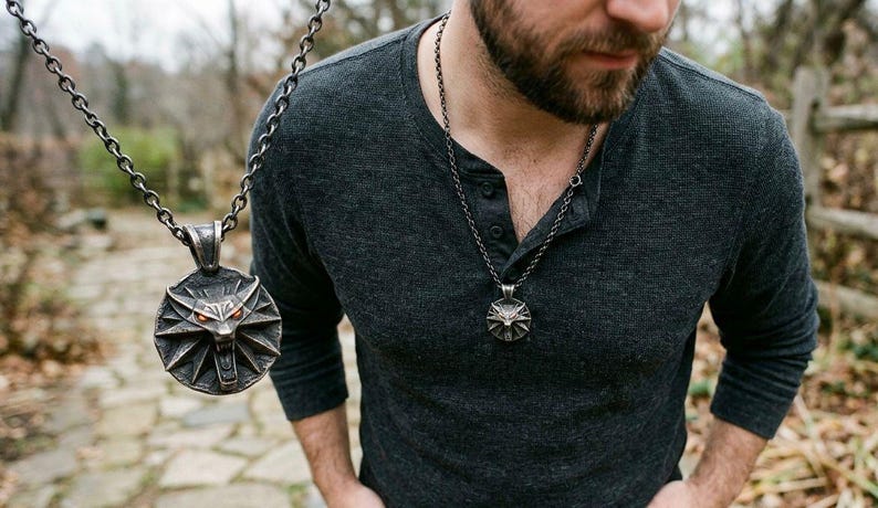 May include: A silver pendant necklace with a wolf's head emblem and red eyes. The circular pendant has a detailed design. The necklace is worn over a dark grey long-sleeved shirt. The background is a blurred outdoor scene.