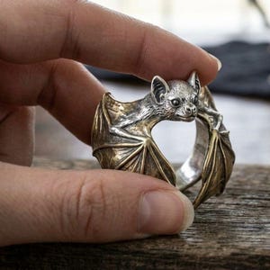 Puede incluir: Un anillo de tono plateado con un diseño detallado de murciélago con las alas extendidas. La cabeza del murciélago está situada en la parte superior central, mientras que las alas envuelven la banda. El metal tiene un acabado pulido con zonas oscuras.
