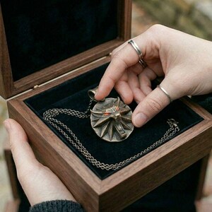 May include: A bronze pendant necklace with a geometric design, displayed in a wooden box with a black velvet lining. The necklace is on a chain. The box is held open by a person.