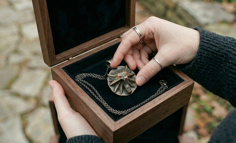 May include: A bronze pendant necklace with a geometric design, displayed in a wooden box with a black velvet lining. The necklace is on a chain. The box is held open by a person.