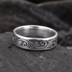 May include: A silver ring with an engraved floral pattern. The ring has a polished finish and a detailed design of swirling leaves and vines. The ring is displayed on a dark, textured surface.