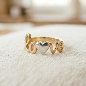 Puede incluir: Un anillo dorado con la palabra "love" escrita en cursiva. Un corazón plateado está centrado entre las letras. El anillo se muestra sobre una superficie de color neutro.