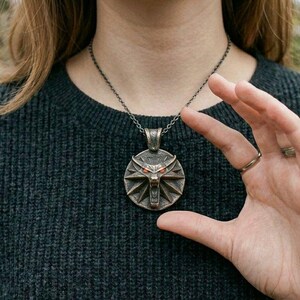 May include: A bronze pendant necklace with a wolf's head emblem and red eyes. The pendant hangs from a dark chain. The person is wearing a dark grey sweater.