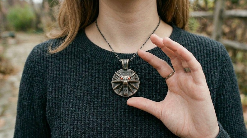 May include: A bronze pendant necklace with a wolf's head emblem and red eyes. The pendant hangs from a dark chain. The person is wearing a dark grey sweater.