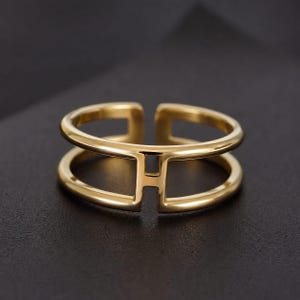 Peut inclure: Une bague dorée au design ouvert. La bague présente deux bandes parallèles reliées par une barre verticale, formant la lettre "H". La bague est posée sur un fond sombre, soulignant sa finition métallique brillante.