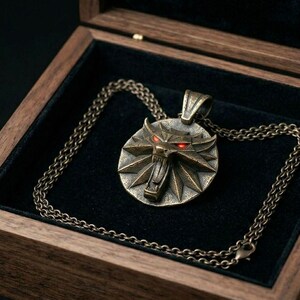 May include: A bronze pendant necklace with a wolf emblem and red eyes, displayed in an open wooden box with a black velvet lining. The chain is also bronze. The box is made of wood.