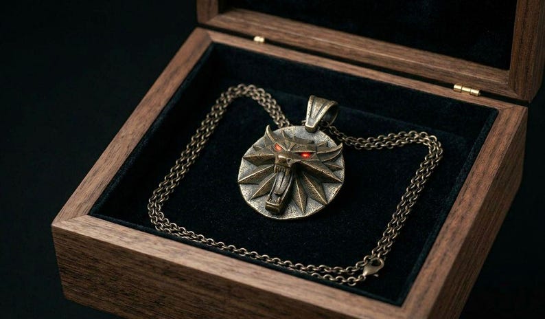 May include: A bronze pendant necklace with a wolf emblem and red eyes, displayed in an open wooden box with a black velvet lining. The chain is also bronze. The box is made of wood.