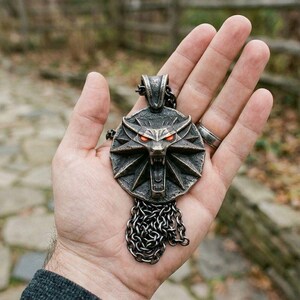 May include: A detailed, antique-style pendant featuring a wolf's head with glowing red eyes. The pendant is made of a dark metal and hangs from a chain. The design is intricate and detailed, with a weathered appearance.