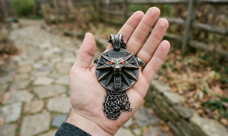 May include: A detailed, antique-style pendant featuring a wolf's head with glowing red eyes. The pendant is made of a dark metal and hangs from a chain. The design is intricate and detailed, with a weathered appearance.
