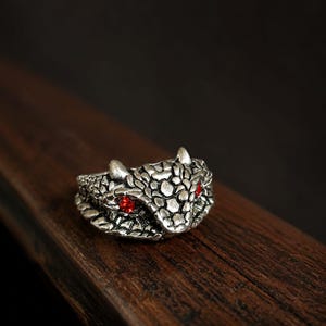May include: A silver-toned metal ring shaped like a snake head with textured scales and two small, round, bright red stones set as eyes. The ring features a detailed, raised pattern across the surface, depicting the head of a serpent.