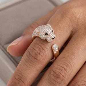May include: A rose gold ring featuring a panther head design. The panther's head and the ring's band are encrusted with small, sparkling clear crystals. The panther's eyes are black. The ring is on a finger.