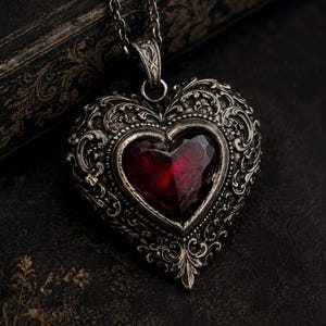 May include: A heart-shaped pendant necklace featuring a deep red faceted gemstone set in an ornate, antiqued silver-toned metal frame with intricate scrollwork patterns, suspended from a matching silver-toned metal chain link necklace.