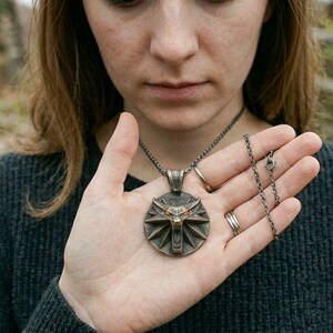 May include: A bronze pendant necklace with a circular wolf head emblem. The necklace is held in a person's hand, highlighting the pendant's details and chain. The person is wearing a dark grey jumper. The background is blurred.