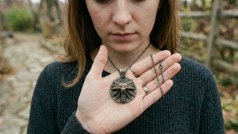May include: A bronze pendant necklace with a circular wolf head emblem. The necklace is held in a person's hand, highlighting the pendant's details and chain. The person is wearing a dark grey jumper. The background is blurred.