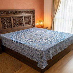 Könnte beinhalten: Ein Holzbett mit geschnitztem Kopfteil ist mit einer blau-weiß gemusterten Mandala-Bettdecke bedeckt. Der Raum hat Holzböden, einen kleinen Teppich und einen Nachttisch mit einer Lampe.