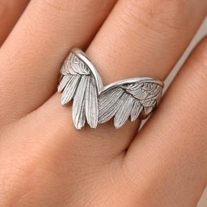 May include: A silver-colored ring shaped like angel wings. The ring features detailed feather textures and is worn on a finger. The wings curve upwards, creating a V-shape. The ring is a piece of jewelry.