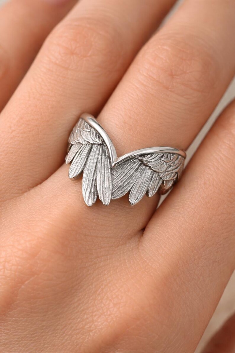 May include: A silver-colored ring shaped like angel wings. The ring features detailed feather textures and is worn on a finger. The wings curve upwards, creating a V-shape. The ring is a piece of jewelry.