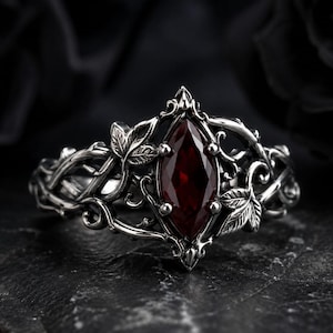 Puede incluir: Un anillo de tono plateado con una piedra preciosa de color rojo intenso en talla marquesa en el centro. La banda presenta un intrincado trabajo en metal que representa enredaderas entrelazadas y hojas detalladas que enmarcan la piedra.