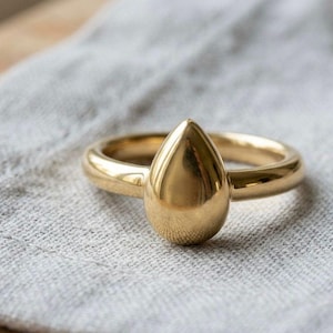 Op de afbeelding: Een goudkleurige ring met een druppelvormige middensteen. De ring ligt op een lichtgrijze linnen doek. De band van de ring is dik en afgerond, en de druppel is glad en gepolijst.