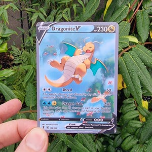 May include: A Dragonite V Pokémon trading card. The card features a cartoon illustration of a beige dragon with blue wings. The card has a blue and gold border and includes text describing the character's abilities and stats.