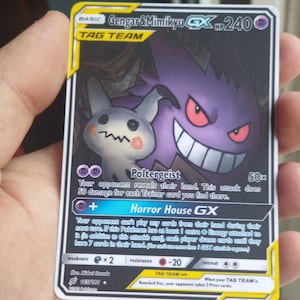 May include: A Pokémon trading card featuring Gengar & Mimikyu GX. The card has a purple and white colour scheme with the text "TAG TEAM" at the top. The card's HP is 240. The card has the text "Poltergeist" and "Horror House GX".
