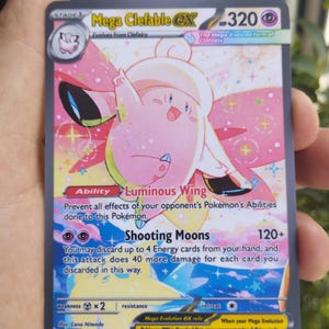 May include: A Mega Clefable EX Pokémon card featuring a pink and white cartoon character with the text "Luminous Wing" and "Shooting Moons". The card has a HP of 320 and includes the text "Mega Evolution EX rule".
