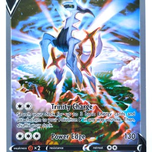 May include: Arceus V Pokémon trading card depicting a white and blue creature with gold detailing. The card displays a HP of 220 and features the text "Trinity Charge" and "Power Edge". The background shows a landscape with mountains and clouds.