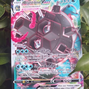 Peut inclure: Une carte à collectionner Pokémon avec Blastoise VMAX. La carte présente un schéma de couleurs bleu et noir avec le texte "Blastoise VMAX" et "Gigantamax". Les noms d'attaque de la carte sont "Grand Falls" et "G-Max Bombard". La carte est entourée de feuilles vertes.